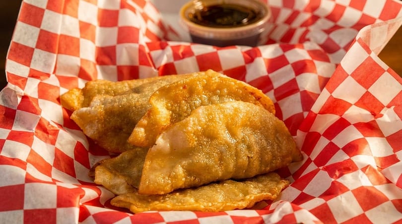Golden-brown fried dumplings in a red and white checkered paper-lined basket with a side of sauce.