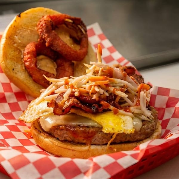 A burger with a fried egg, bacon, coleslaw, and onion rings on a red and white checkered liner.