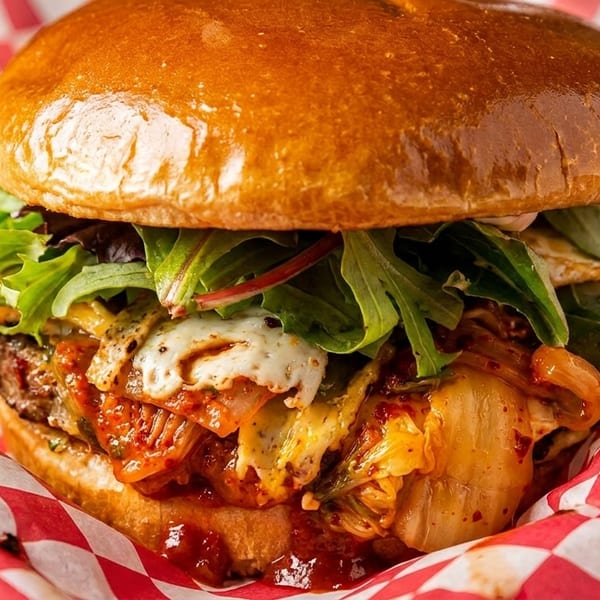 A juicy burger with kimchi, melted cheese, and fresh greens, served in red and white checkered paper.