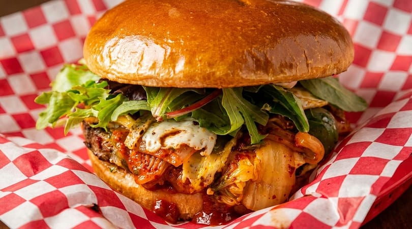 A juicy burger with kimchi, melted cheese, and fresh greens, served in red and white checkered paper.