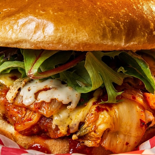 Close-up of a burger with kimchi, melted cheese, and lettuce, served in red and white checkered paper.